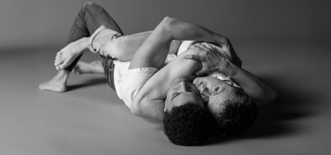 Kaksi miestä makaa lattialla intiimissä, läheisessä syleilyssä vastakkain. Heillä on rennot vaatteet ja he näyttävät jakavan hellän hetken. Kuva on mustavalkoinen, mikä korostaa heidän yhteyttään.