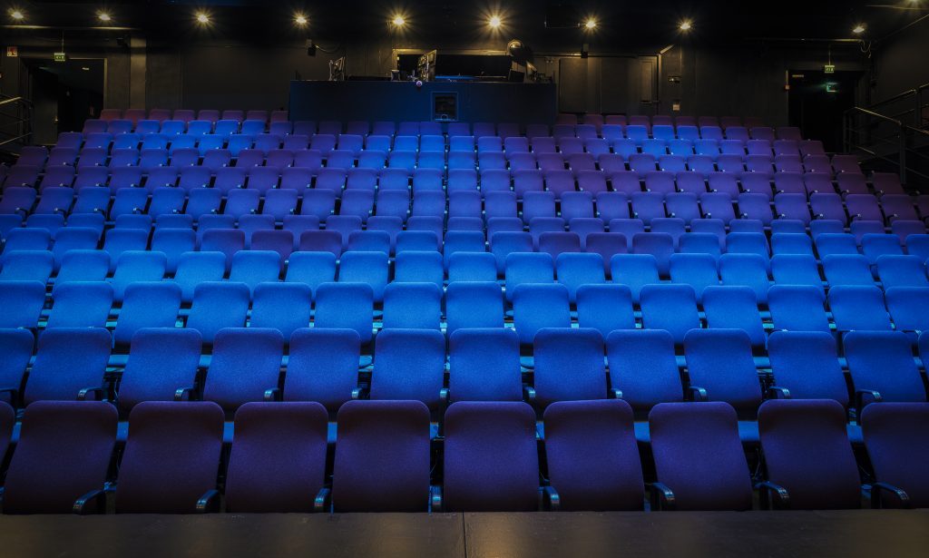 Tyhjä teatterisali, jossa on sinisiä ja violetteja penkkirivejä, joiden takana on ohjauskoppi, jota valaisevat pimeässä huoneessa yläpuolella olevat valonheittimet.