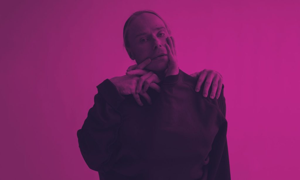 Tummapaitainen henkilö seisoo vaaleanpunaista taustaa vasten, ja toisen henkilön kädet koskettavat hellästi hänen kasvojaan ja olkapäätään, mikä luo surrealistisen vaikutelman.