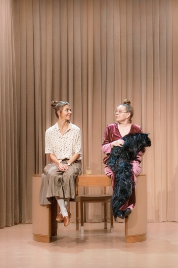 Kaksi naista istuu penkillä beigen verhojen edessä. Toisella on pusero ja hame, toisella on silmälasit ja vaaleanpunainen asu, ja hänellä on kädessään suuri musta karvainen esine. He katsovat toisiaan. Heidän välissään istuu vesilasi.