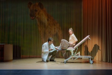 Lääkäri polvistuu tutkimuspöydällä istuvan naisen viereen; heidän takanaan näyttämöverhoihin heijastetaan suuri hyeenan kuva, joka luo surrealistisen tunnelman.