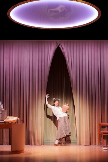 Eläimen korviin ja häntään pukeutunut esiintyjä poseeraa dramaattisesti pitäen kädessään suurta mukia. Hän seisoo lilojen verhojen edessä kirkkaasti valaistulla näyttämöllä, jonka yläpuolella on pyöreä valo.