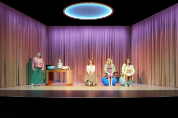 Neljä naista istuu ja seisoo lavalla, jonka yläpuolella on violetit verhot ja hehkuva pyöreä valo. Kolme naista istuu jumppapallojen päällä; yksi nainen seisoo pöydän vieressä, jossa on purkkeja ja kulhoja. Kohtaus on kirkkaasti valaistu, ja siinä on pastellisävyjä.