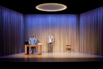 Kaksi ihmistä on näyttämöllä, jossa on suljetut beiget verhot ja pyöreä kattovalaisin. Toinen seisoo pöydän takana kannettavan tietokoneen kanssa, kun taas toinen seisoo pitelemässä esinettä rintansa edessä. Oikealla puolella on tuoli.