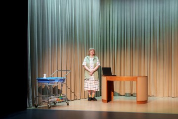 Henkilö seisoo lavalla puisen työpöydän vieressä, jossa on suljettu kannettava tietokone ja ostoskärry täynnä siivoustarvikkeita, korkeiden beigen verhojen edessä.