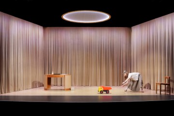 Pukuun pukeutunut henkilö kumartuu työntämään suurta leluautoa kirkkaasti valaistulla näyttämöllä, jossa on beiget verhot, pyöreä valo yläpuolella ja minimaaliset huonekalut, kuten pöytä ja tuoli.