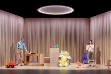 Lavastuksessa vasemmalla on mies työpöydän ääressä ja oikealla eläinkorvainen nainen, jolla on oranssi paita kädessään. Taustalla on beiget verhot, ja näyttämö on koristeltu suurilla leluilla ja lastenkalusteilla.