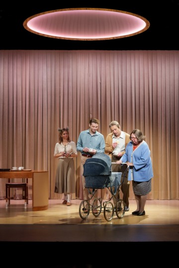 Neljä aikuista seisoo lavalla sinisten vanhojen lastenvaunujen ympärillä pyöreän valon alla. Heidän takanaan on beigeistä pystysuorista verhoista koostuva yksinkertainen tausta ja puinen pöytä, jossa on teetarvikkeita. He näyttävät keskittyvän vaunuihin.