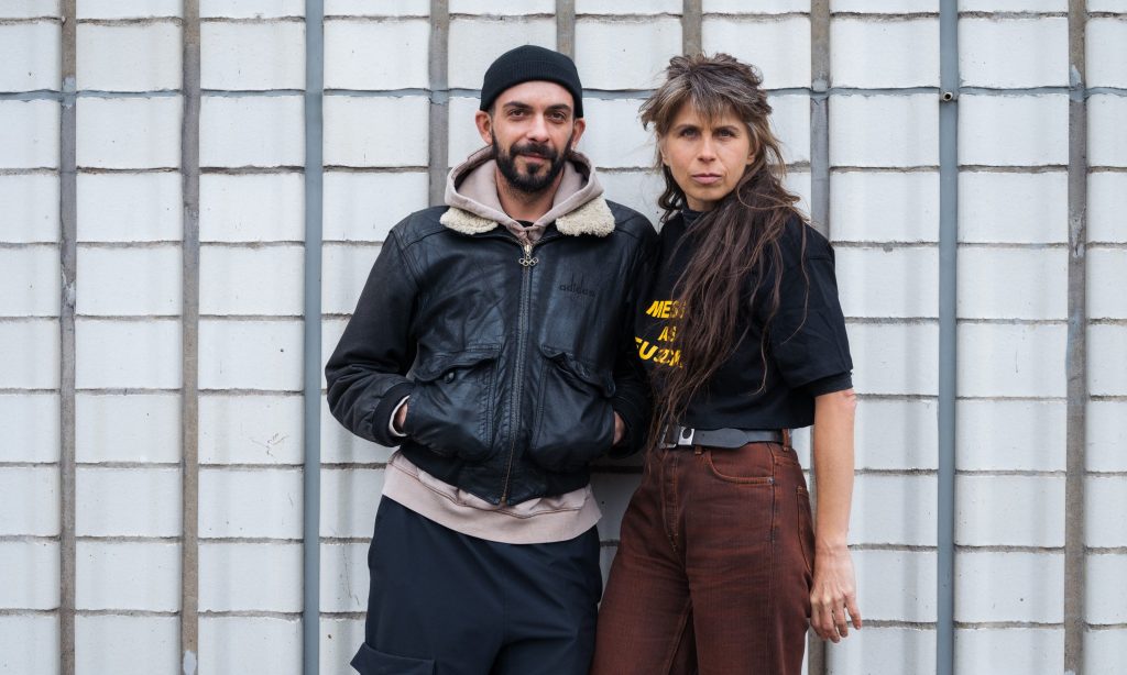 Kaksi ihmistä seisoo vierekkäin valkoisen tiiliseinän edessä. Vasemmalla olevalla henkilöllä on musta pipo, musta takki ja tummat housut. Oikealla olevalla henkilöllä on pitkät hiukset, hänellä on yllään musta t-paita ja ruskeat housut.