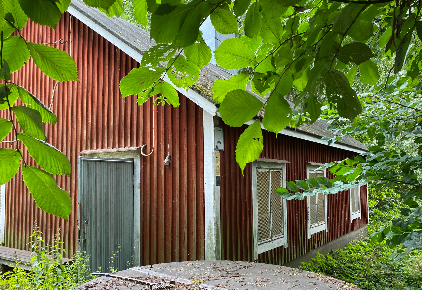 Punainen puurakennus, jossa on harmaa ovi ja useita valkoisiksi kehystettyjä ikkunoita, on osittain rehevien vihreiden lehtien ja puiden peitossa.