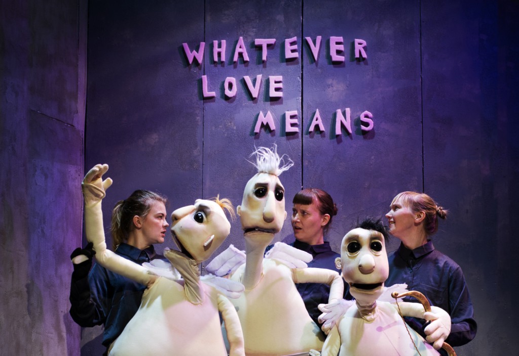 Neljä tummiin vaatteisiin pukeutunutta nukketeatteritaiteilijaa pitelee suuria valkoisia nukkeja violetin seinän edessä, jonka yläpuolella on vaaleanpunaisin kirjaimin kirjoitettu teksti WHATEVER LOVE MEANS.
