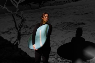 Kylpypukuun pukeutunut nainen pitää surffilautaa edessään ja seisoo tummaa, surrealistista taustaa vasten, jossa on abstrakteja varjoja ja karu puu. Hänen ilmeensä on vakava ja mietteliäs.