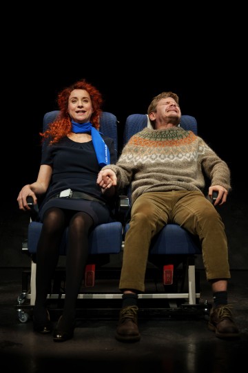 Siniseen mekkoon ja huiviin pukeutunut nainen pitää kädestä kiinni kuviopuseroon pukeutuneen miehen kanssa, kun he istuvat lavalla sinisissä lentokoneen istuimissa. Mies vaikuttaa jännittyneeltä, kun taas nainen on rauhallinen. Tausta on tumma.