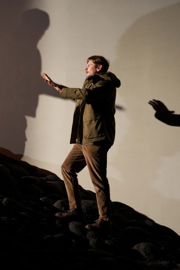 Vihreään takkiin pukeutunut mies seisoo epätasaisilla kivillä, kurottaa molemmilla käsillään eteenpäin ja heittää suuren varjon kalpealle seinälle takanaan. Toinen osittainen varjo ilmestyy seinälle oikealla.