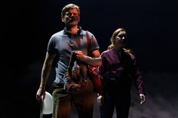 Mies, jolla on kädessään tuoli ja karvainen esine, seisoo dramaattisessa valaistuksessa naisen vieressä, jolla on kaulanauha, ja molemmat katsovat eteenpäin tummaa, savuista taustaa vasten.