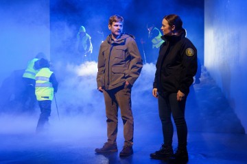 Kaksi ihmistä seisoo sinisen valon ja sumun alla ulkoilutakit yllään, kun taas heijastinliiveihin ja huppuihin pukeutuneet hahmot työskentelevät sumuisessa taustassa. Kohtaus vaikuttaa dramaattiselta ja jännittyneeltä.