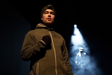 Pipoon ja vetoketjulliseen takkiin pukeutunut mies seisoo pimeässä huoneessa käsi rinnalla. Hänellä on otsalamppu, ja valonheitin valaisee taustalla olevaa varjoista hahmoa, mikä luo dramaattisen sumuisen kohtauksen.