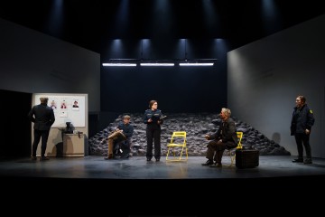 Viisi ihmistä on hämärästi valaistulla näyttämöllä, jonka taustalla on kivinen tausta. Kolme istuu tai seisoo keltaisten tuolien ääressä, yksi valokuvia sisältävän taulun vieressä ja yksi seisoo erillään. Kohtaus herättää jännittyneen tai tutkivan ilmapiirin.