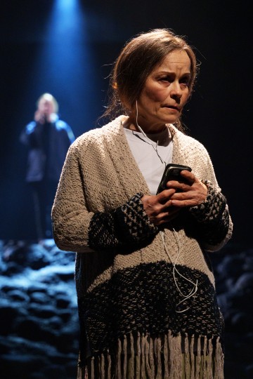 Nainen seisoo valonheittimen alla, kädessään älypuhelin, jossa on kuulokkeet, ja näyttää mietteliäältä. Taustalla on hämärä hahmo, joka seisoo toisen valonheittimen alla tummalla, kuvioidulla näyttämöllä.