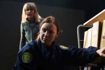 Kaksi naista sisätiloissa: toinen etualalla tummassa poliisin univormussa ja vakavana kurkottaen kansioihin; toinen nainen seisoo hänen takanaan varjossa, osittain epäselvästi.