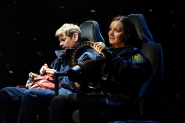 Kaksi poliiseiksi pukeutunutta ihmistä istuu lavalla autonistuimissa. Oikeanpuoleinen nainen pitelee ratista ja näyttää ajavan, kun taas vasemmanpuoleinen mies katsoo alaspäin keskittyneenä johonkin käsissään olevaan esineeseen.