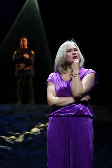 Harmaahiuksinen nainen violetissa asussaan seisoo mietteliäänä käsi leualla valonheittimen alla, kun taas toinen hahmo seisoo epäselvästi taustalla valonsäde heidän yläpuolellaan.