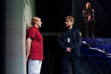 Punaisiin leikkausasuihin pukeutunut henkilö nojaa seinää vasten ja puhuu turvapukuun pukeutuneelle henkilölle. Toinen virkapukuinen henkilö seisoo heidän takanaan kivisellä, hämärästi valaistulla lavalla.