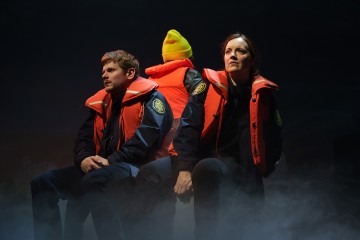 Kaksi oransseihin pelastusliiveihin pukeutunutta ihmistä istuu selkä toisensa perään sumuisella näyttämöllä, ja molemmilla on yllään tummat univormut, joissa on laastareita. Keltaiseen pipoon ja pelastusliiviin pukeutunut henkilö istuu heidän välissään, kasvot poispäin. Kohtaus on hämärästi valaistu ja dramaattinen.