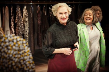Kaksi hymyilevää vanhempaa naista seisoo vaatekaupassa, toinen mustassa topissa ja ruskeankeltaisessa hameessa, toinen vaaleanvihreässä kuvioidussa takissa, ja taustalla näkyy vaatehyllyjä ja toinen henkilö.