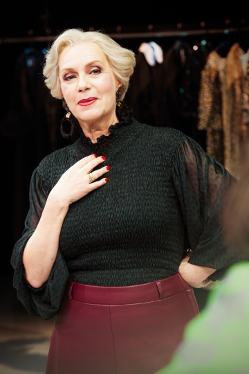 Elegantti vanhempi nainen, jolla on lyhyet, muotoillut vaaleat hiukset, tumma pusero ja ruskeankeltainen hame, seisoo sisätiloissa käsi rinnalla ja katsoo mietteliäästi sivulle. Vaatehyllyt näkyvät epäselvässä taustassa.