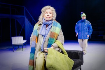 Lavalla seisoo vanhempi kiharatukkainen nainen, jolla on värikäs huivi ja talvitakki, ja hänellä on kädessään vihreä huopa. Taustalla kävelee siniseen talvitakkiin ja pipoon pukeutunut mies. Lavastus on minimaalisesti kalustettu ja hämärästi valaistu.