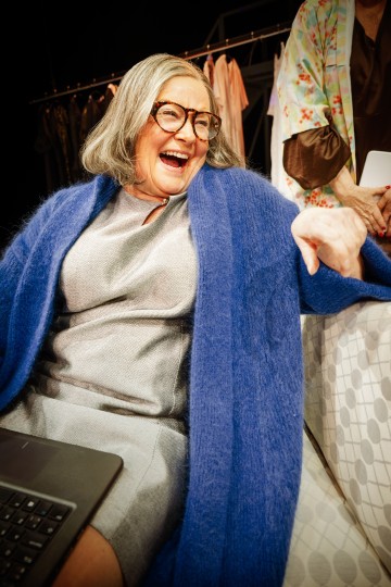 Vanhempi nainen, jolla on harmaat hiukset ja silmälasit, harmaa mekko ja sininen neuletakki, nauraa iloisesti istuen kuviollisella sohvalla. Hänen takanaan olevassa telineessä on vaatteita, ja lähellä on kannettava tietokone.