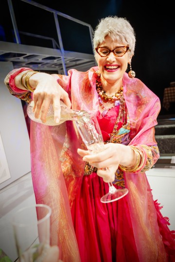 Vanhempi nainen, jolla on lyhyet harmaat hiukset, silmälasit ja värikkäitä koruja, kaataa iloisesti juomaa lasiin yllään kirkkaan vaaleanpunainen mekko ja huivi. Hän hymyilee ja näyttää juhlivan.