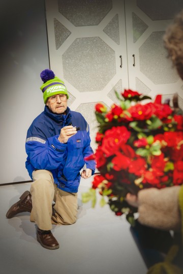 Siniseen takkiin ja vihreään talvihattuun pukeutunut iäkäs henkilö polvistuu sisätiloissa pienen laatikon kanssa. Etualalla on epäselvästi suuri punaisia ruusuja sisältävä kimppu.