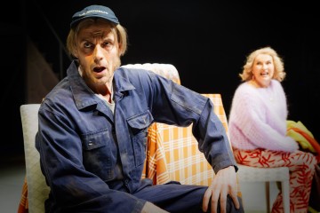 Siniseen haalariin ja lippikseen pukeutunut mies istuu pöydän ääressä, jossa on oranssiruutuinen pöytäliina, ja näyttää yllättyneeltä. Vaaleanpunaiseen puseroon ja punakuvioisiin housuihin pukeutunut nainen istuu hänen takanaan ja hymyilee. Molemmat näyttävät olevan lavalla.