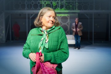 Vihreään villapaitaan ja huiviin pukeutunut harmaahiuksinen nainen pitelee vaaleanpunaista laukkua kirkkaiden valojen alla lavalla. Taustalla näkyy kaksi epäselvää hahmoa, mikä viittaa teatteriesitykseen.