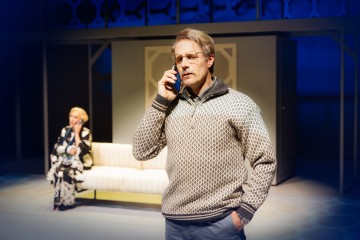 Silmälasipäinen ja kuviopuseroon pukeutunut mies puhuu kännykkään ja näyttää vakavalta. Taustalla istuu nainen valkoisella sohvalla, joka myös puhuu puhelimeen. Kohtaus näyttää olevan lavastettu näyttämölle, jossa on dramaattinen valaistus.