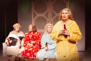 Neljä värikkäisiin kylpytakkeihin pukeutunutta naista istuu sohvalla, ja jokaisella on kädessään olutpullo. Kolme istuu yhdessä taustalla, kun taas yksi seisoo edessä puhumassa tai esiintymässä keltaiseen kaapuun pukeutuneena. Asetelma vaikuttaa teatraaliselta.