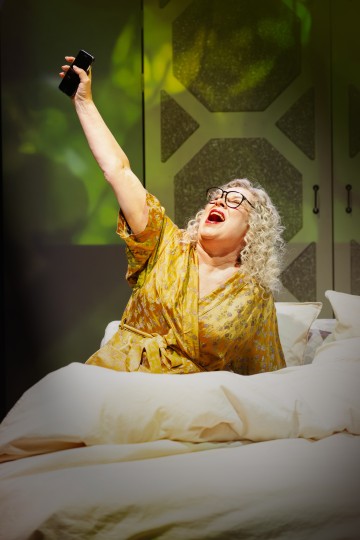 Vanhempi nainen, jolla on kiharat harmaat hiukset ja silmälasit, istuu sängyssä, nauraa iloisesti, on pukeutunut keltaiseen kylpytakkiin ja pitää toisessa kädessään television kaukosäädintä. Tausta on lämpimästi valaistu kelta-vihreillä sävyillä.