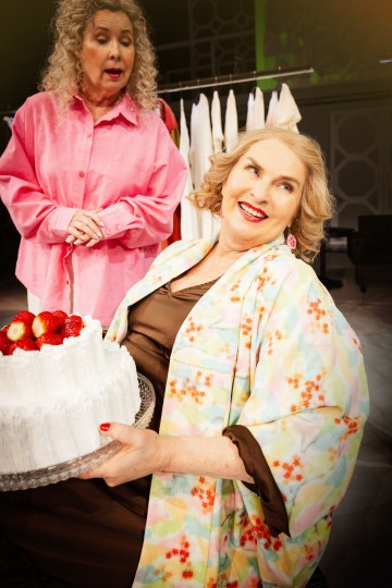 Kaksi vanhempaa naista sisätiloissa; toinen värikkääseen kaapuun pukeutunut nainen hymyilee pitäen kädessään valkoista mansikoilla kuorrutettua kakkua, ja toinen vaaleanpunaiseen paitaan pukeutunut nainen seisoo puhuessaan hänen takanaan, ja taustalla on roikkuvia valkoisia vaatteita.