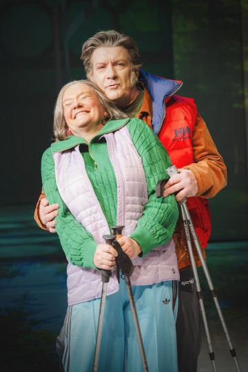 Kaksi iäkästä aikuista hymyilee syleillessään toisiaan. Molemmat ovat pukeutuneet värikkäisiin ulkoiluvaatteisiin ja pitävät kädessään kävelysauvoja, mikä viittaa siihen, että he ovat vaeltamassa tai nauttimassa kävelystä yhdessä.