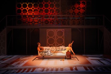 Kaksi näyttelijää esiintyy näyttämöllä; toinen makaa sängyssä peiton alla, kun taas toinen istuu sängyn juurella ja osoittaa ylöspäin. Taustalla on geometrisia kuvioita ja dramaattinen puna-oranssi valaistus.