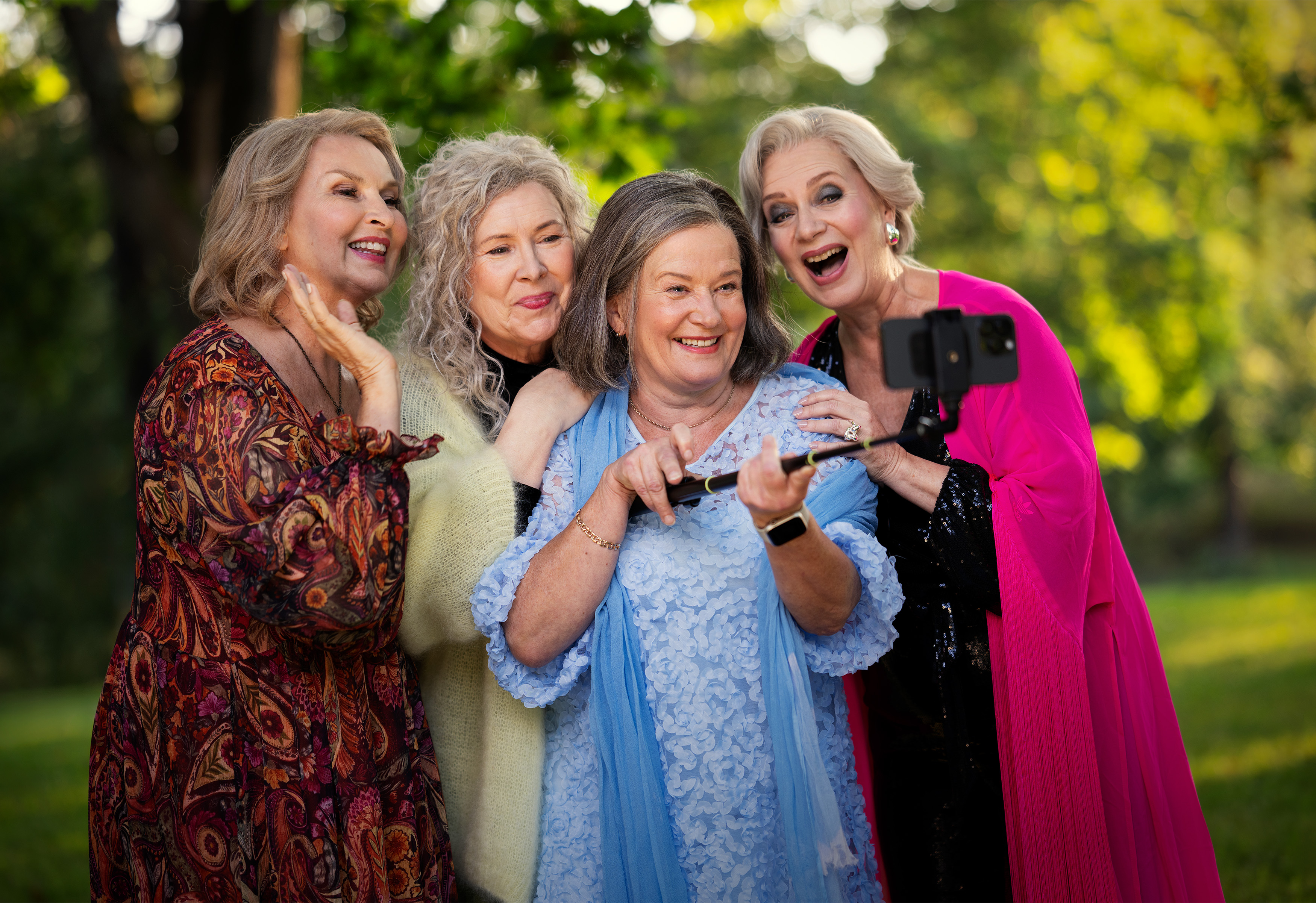 Neljä vanhempaa naista seisoo lähekkäin ulkona, hymyilee ja poseeraa selfietä varten älypuhelimella selfiekepin varassa. He ovat pukeutuneet kirkkaisiin, värikkäisiin vaatteisiin, ja tausta on vihreä ja vehreä.