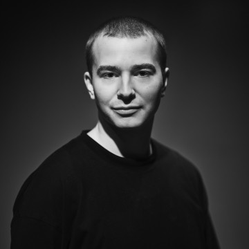 Lyhythiuksinen nuori mies seisoo tumman taustan edessä, päällään musta paita. Hän katsoo kameraan hymyillen, ja dramaattinen valaistus korostaa hänen kasvojaan. Kuva on mustavalkoinen.