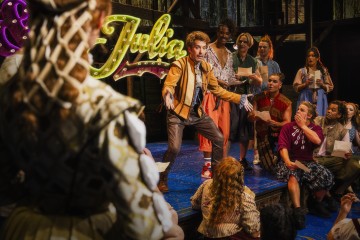Vilkkaassa näyttämökohtauksessa esiintyjä, jolla on yllään ruskea takki, laulaa pukeutuneelle ryhmälle, ja taustalla näkyy hehkuva kyltti, jossa lukee ”Juliet”. Yleisö on mukana esityksessä ja ilmaisee tunteitaan eloisasti.