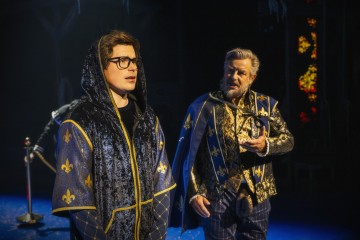 Kaksi näyttelijää lavalla on pukeutunut keskiaikaisiin kaapuihin, joissa on kultaisia liljakuvioita. Toinen heistä, nuorempi ja silmälasipäinen, seisoo etualalla, kun taas vanhempi näyttelijä elehtii ilmeikkäästi taustalla. Näyttämö on hämärästi valaistu.