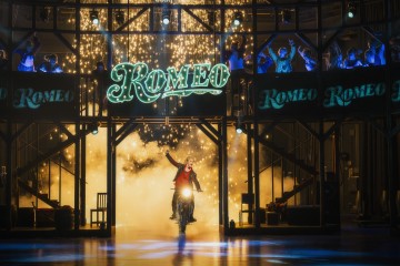 Esiintyjä ajaa moottoripyörällä lavalla kirkkaan ROMEO-kyltin alla, kun kipinöitä sataa ja näyttelijät hurraavat hänen yläpuolellaan ja takanaan.