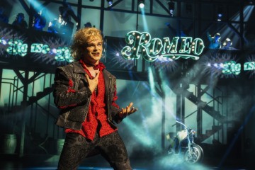 Näyttämöpuvussa oleva esiintyjä laulaa lavalla, kun taustalla näkyy neonkyltti, jossa lukee ”ROMEO”, sekä moottoripyörä, ja heitä valaisee värikäs teatterivalaistus.