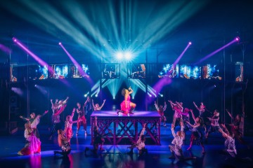 Teatterilavalla, jota dramaattinen valaistus korostaa, näkyy vaaleanpunaisiin pukeutunut esiintyjä korokkeella, jota ympäröivät dynaamisissa asennoissa olevat tanssijat, ja siniset ja violetit kohdevalot valaisevat kohtausta.