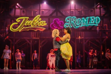 Kaksi näyttelijää syleilee toisiaan näyttämön keskellä Julia & Romeo -neonkyltien alla, ja taustalla näkyvät muut näyttelijät sekä dramaattinen näyttämövalaistus, mikä luo eloisan teatterikohtauksen.